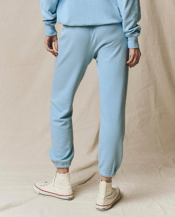 A person stands on a light fabric surface, photographed from the back, wearing THE GREAT. SU25 SEASONAL KNITS’ The Stadium Sweatpant. Solid -- Vista Blue with a matching sweatshirt and white high-top sneakers.