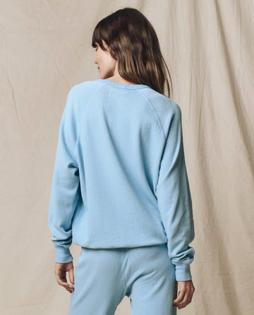 A person with long brown hair, wearing THE GREAT.'s The College Sweatshirt in Solid Vista Blue from the SU25 SEASONAL KNITS collection, stands facing away from the camera against a beige fabric background.