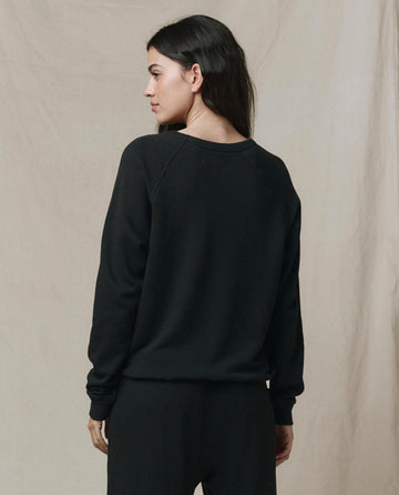 A person with long dark hair faces away, wearing SP26 New Knits' The Rookie Sweatshirt in Solid -- Almost Black and black pants against a plain beige background.
