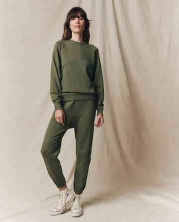 A woman stands before a beige fabric backdrop wearing THE GREAT. SU25 SEASONAL KNITS The Stadium Sweatpant. Solid in Heritage Army with a matching sweatshirt and white high-top sneakers.