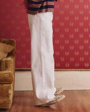 A person in THE GREAT. SP26 D1 The Slouch Wide Leg Jean in True White and sneakers stands beside a mustard sofa, with red patterned wallpaper in the background.