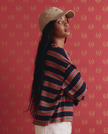 A woman with long black hair wears THE GREAT. SP26 D1’s The Field Tee in Scout Stripe, paired with a beige baseball cap, standing before a red patterned background.