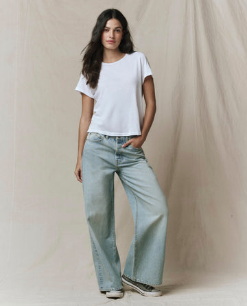 A woman stands against a beige fabric backdrop wearing THE GREAT.'s The Little Tee in True White, inspired by vintage men's undershirts, paired with light blue wide-leg jeans and white sneakers.