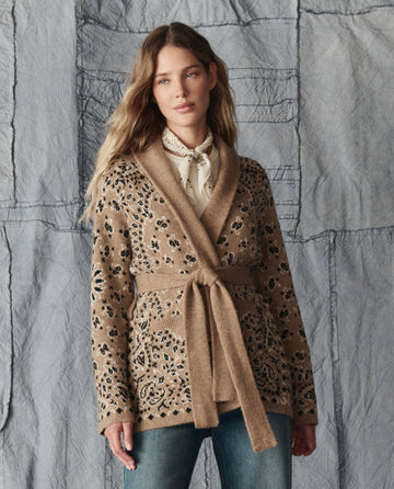 A woman wears THE GREAT. H25 COLLECTION's The Bandana Riding Cardigan in Camel, featuring black floral patterns, over a white blouse and blue jeans, standing before a textured grey fabric backdrop.