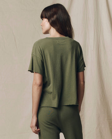 A woman with brown hair faces away, wearing THE GREAT.'s The Crop Tee. Solid in Heritage Army—stylish casual wear from the SU25 SEASONAL KNITS collection—set against a neutral fabric backdrop.