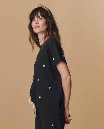 A woman with wavy brown hair wears THE GREAT. PF25 EMB KNITS "The Boxy Crew. Embroidered -- Almost Black With Daisy Embroidery" shirt and matching pants, standing against a beige background.