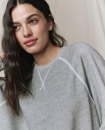 A woman with long dark hair wears THE GREAT. CORE KNITS The Slouch Sweatshirt in Varsity Grey, featuring a straight shape and fuller sleeves, while standing before a neutral background and gazing slightly to the side.