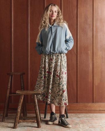 Wearing THE GREAT. SP26 D1 The Trainer Jacket in Robin Blue, a person with long wavy hair, floral skirt, and black sneakers stands indoors by two wooden stools against a wood-paneled wall.