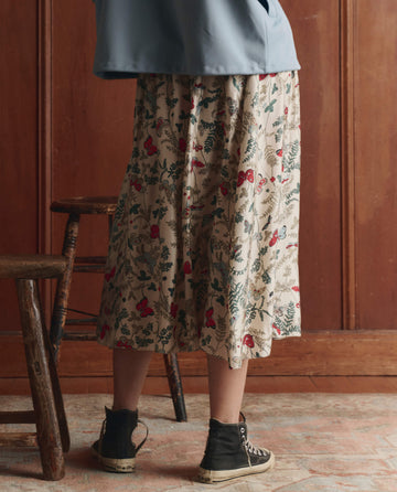 A person wearing THE GREAT. SP26 D1 The Canary Skirt – Floating Butterfly stands in beige, paired with black high-top sneakers and a blue top near wooden chairs against a wood-paneled wall.