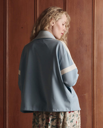 A person with wavy blonde hair wears THE GREAT. SP26 D1 The Trainer Jacket in Robin Blue, featuring white stripes, while standing in front of wooden paneling facing away and looking downward.