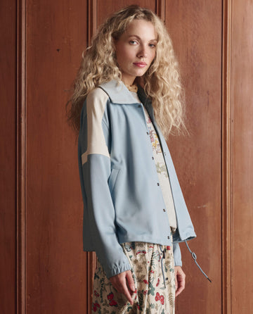 A woman with long curly blonde hair wears THE GREAT. SP26 D1 The Trainer Jacket in Robin Blue over a floral dress, standing before a wooden paneled wall.