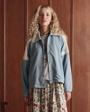 Wearing THE GREAT. SP26 D1's The Canary Skirt — Floating Butterfly, a person with long curly blond hair and a light blue jacket stands before a wood-paneled wall.
