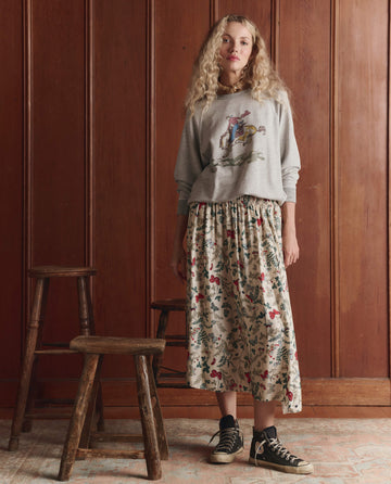 A woman stands indoors in THE GREAT. SP26 D1 The College Sweatshirt w/ Rider Graphic, Novelty—Soft Heather Grey, paired with a floral midi skirt and black sneakers; two wooden stools sit beside her against a wood-paneled wall.
