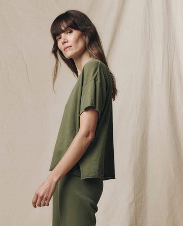 A person with long brown hair stands before a beige fabric backdrop wearing THE GREAT.'s The Crop Tee. Solid in Heritage Army from the SU25 SEASONAL KNITS collection, showcasing effortless casual style.