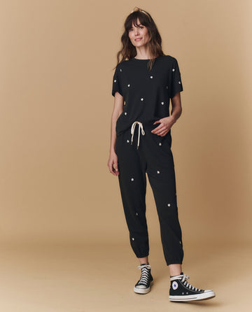 A woman stands against a beige background wearing THE GREAT. PF25 EMB KNITS The Boxy Crew. Embroidered in Almost Black with Daisy Embroidery, paired with jogger pants and black Converse sneakers.
