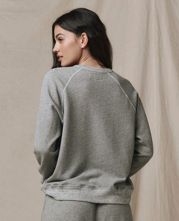 A woman with long dark hair, seen from behind, wears the SP26 New Knits "The Rookie Sweatshirt. Solid -- Varsity Grey" and matching pants, standing before a beige fabric backdrop.