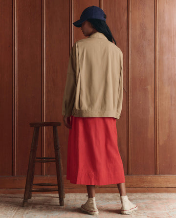 A person stands indoors facing away, wearing THE GREAT. SP26 D1 The Folsom Jacket in Khaki, a red skirt, beige boots, and a navy cap near a wooden stool against a wood-paneled wall.