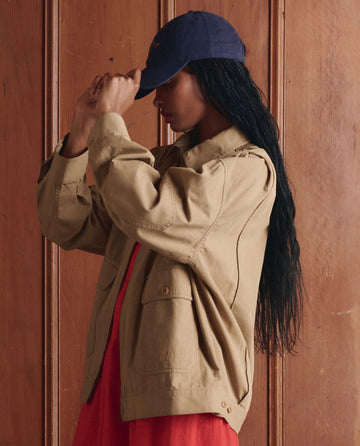 A person wearing THE GREAT. SP26 D1 The Folsom Jacket in Khaki and a navy cap stands before a wooden wall, head slightly lowered and one hand adjusting the cap.