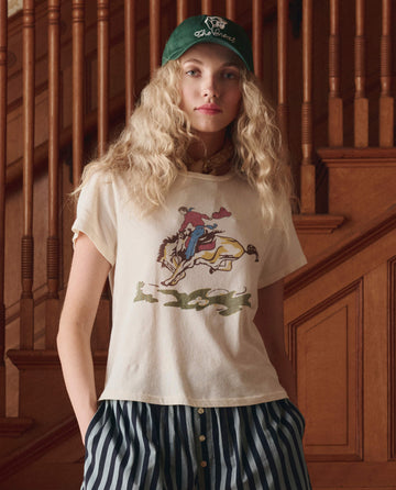 A person with long curly hair, wearing a green cap and THE GREAT. SP26 D1 The Little Tee W/ Rider Graphic in Washed White, stands by a wooden staircase.