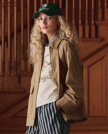 Wearing THE GREAT. SP26 D1 The Little Tee W/ Rider Graphic in washed white, a person with long curly hair stands in front of a wooden staircase, paired with a green cap, tan jacket, and striped pants.