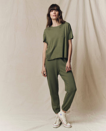A woman stands against a beige backdrop, modeling THE GREAT. SU25 SEASONAL KNITS The Crop Tee. Solid in Heritage Army with matching joggers and white high-top sneakers.