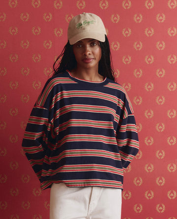 A woman in THE GREAT.'s The Field Tee -- Scout Stripe and white pants stands against a red patterned background.
