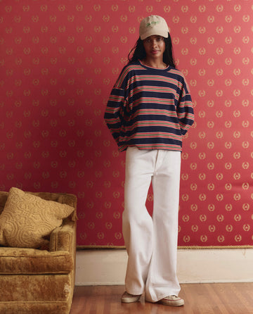 Wearing THE GREAT.'s The Field Tee in Scout Stripe, a person stands by a red patterned wall near a brown couch with a yellow cushion.