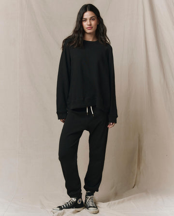 A woman stands before a beige backdrop wearing THE GREAT. CORE KNITS The Slouch Sweatshirt in Almost Black, paired with matching black sweatpants and black Converse sneakers.