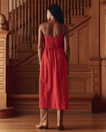 A woman wearing THE GREAT. SP26 D1 The Verse Dress in Hot Red and brown boots stands with her back to the camera in front of a wooden staircase.