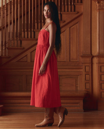 A woman wears THE GREAT. SP26 D1 The Verse Dress in Hot Red with brown cowboy boots, standing indoors by a wooden staircase and paneled walls.