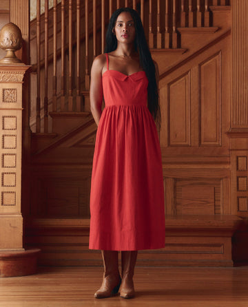 A woman stands indoors in front of a wooden staircase, wearing THE GREAT. SP26 D1 The Verse Dress in Hot Red and brown boots, with her hands behind her back.