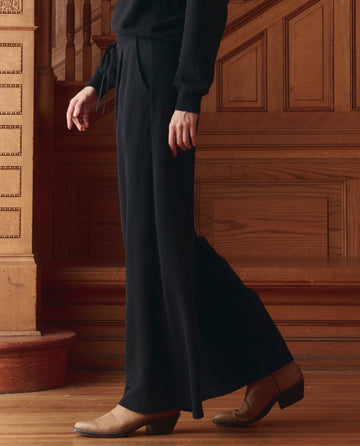 A person wearing THE GREAT. SP26 D1 The Relaxed Trouser Pant in black and tan ankle boots stands before ornate wooden paneling and stairs.
