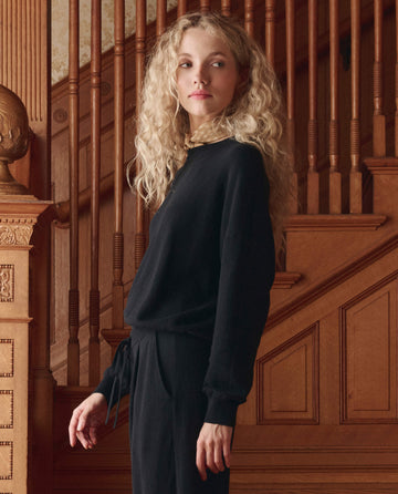A woman with long curly blonde hair wears THE GREAT. SP26 D1 The Oversized Pullover in black, standing before a wooden staircase and glancing over her shoulder.