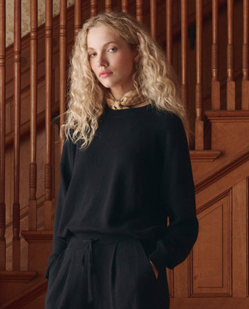 A person with long curly blonde hair wears THE GREAT. SP26 D1 The Oversized Pullover in black, paired with pants, standing before a wooden staircase and looking at the camera.