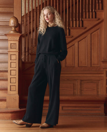 A person with long curly hair wears THE GREAT.'s The Relaxed Trouser Pant in black, paired with brown shoes, standing before a wooden staircase and wall paneling.