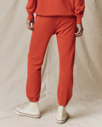 A person stands on a beige fabric backdrop wearing THE GREAT.'s The Stadium Sweatpant. Solid in Desert Clay from the SU25 SEASONAL KNITS, paired with a matching orange top and white high-top sneakers.