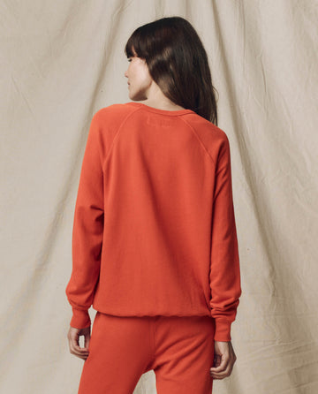 A person with long hair stands with their back to the camera, wearing THE GREAT. SU25 SEASONAL KNITS The College Sweatshirt in Solid Desert Clay, set against a beige fabric backdrop.