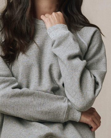 A person with long brown hair models the SP26 New Knits The Rookie Sweatshirt in Varsity Grey, posing with one hand on their chin and the other arm crossed. Their face is partially out of frame.