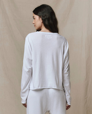A woman with long dark hair faces away, wearing THE GREAT. CORE KNITS The Long Sleeve Crop Tee in True White and matching white pants against a beige background.