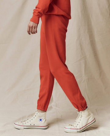 A person models THE GREAT.'s The Stadium Sweatpant. Solid in Desert Clay, from the SU25 SEASONAL KNITS collection, walking against a beige stadium backdrop—only their lower body and white Converse sneakers are visible.
