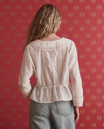 A person with long, wavy blonde hair faces a red patterned wall wearing THE GREAT. SP26 D1 Harper Top in True White—a women’s long-sleeve top with lace details—and light blue jeans.