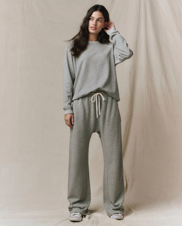 A person stands against a neutral fabric backdrop, wearing the SP26 New Knits The Rookie Sweatshirt in Varsity Grey with matching sweatpants and white sneakers.