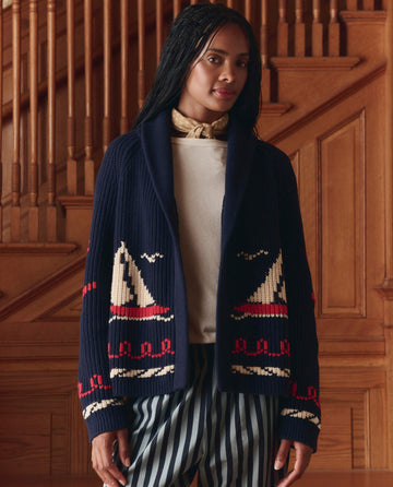 A woman stands indoors by a wooden staircase, wearing THE GREAT. SP25 COLLECTORS The Sailboat Lodge Cardigan in Dark Navy—a hand-embroidered cotton blend sweater with sailboat motifs—over a light shirt, striped pants, and a scarf.