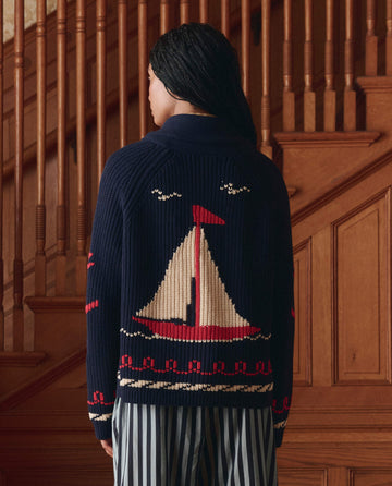 A person stands indoors facing away, wearing THE GREAT. SP25 COLLECTORS The Sailboat Lodge Cardigan in Dark Navy with hand-embroidered boats and seagulls on the back, paired with striped pants. Wood-paneled stairs are behind them.