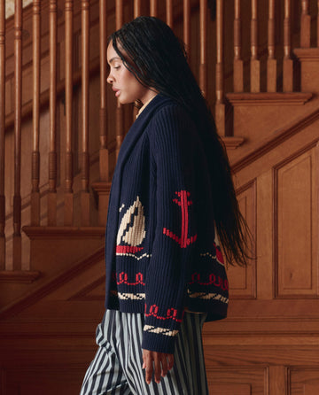 A woman with long braided hair wears THE GREAT. SP25 COLLECTORS The Sailboat Lodge Cardigan in Dark Navy, featuring nautical designs and striped pants, standing before a wooden staircase.
