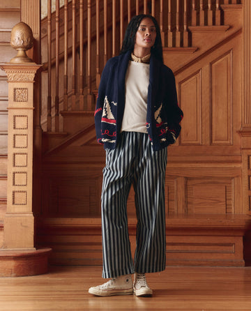 Indoors before a wooden staircase, a person wears THE GREAT. SP25 COLLECTORS The Sailboat Lodge Cardigan in Dark Navy with hand-embroidered boats, paired with a white shirt, striped wide-leg pants, and white high-top sneakers.