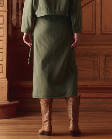 Wearing THE GREAT.'s The Wrap Skirt in Classic Army, a person stands indoors in brown cowboy boots and a green cotton herringbone jacket, highlighting versatile styling against wood-paneled walls and stairs.