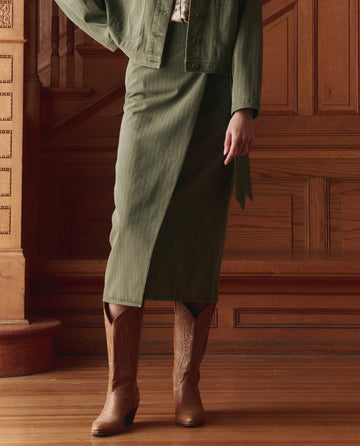 A person wearing THE GREAT. SP26 D1 The Wrap Skirt -- Classic Army, paired with a matching jacket and brown cowboy boots, stands indoors on a wooden floor with wood paneling—an ideal look for versatile styling.