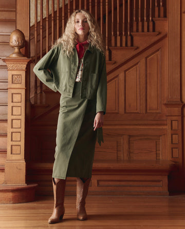 A woman stands by a wooden staircase wearing THE GREAT. SP26 D1's The Wrap Skirt in Classic Army, styled with a green cotton herringbone jacket, red scarf, and brown cowboy boots for a versatile look.