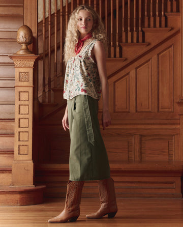 A woman with curly hair stands indoors by a wooden staircase, styled in THE GREAT. SP26 D1’s The Wrap Skirt – Classic Army, teamed with a sleeveless floral blouse and brown cowboy boots for versatile flair.